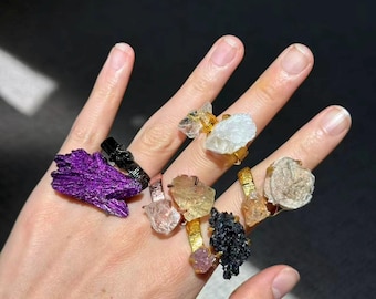 Könnte beinhalten: Eine Hand, die eine Sammlung von Edelsteinringen zeigt. Die Ringe weisen verschiedene rohe, ungeschliffene Steine in Farben wie Lila, Schwarz, Klar und Gold auf, die auf Gold- oder Silberbändern gefasst sind. Die Ringe sind in verschiedenen Größen und Formen.
