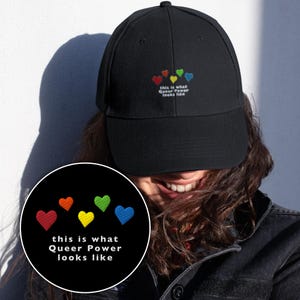Könnte beinhalten: Schwarze Baseballkappe mit gestickten Regenbogenherzen und dem Text "this is what Queer Power looks like." Die Kappe hat einen gebogenen Schirm und ein strukturiertes Design. Die Stickerei umfasst rote, orange, gelbe, grüne und blaue Herzen.