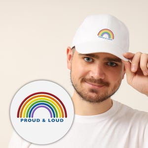 Könnte beinhalten: Weiße Baseballkappe mit Regenbogen-Design und dem Text "PROUD & LOUD". Der Regenbogen hat die Farben Rot, Orange, Gelb, Grün, Blau und Lila. Die Kappe ist aus einem weichen Material gefertigt.