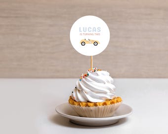 Könnte beinhalten: Ein weißer Cupcake mit gelben und weißen Streuseln, bedeckt mit einem weißen Frosting-Wirbel. Ein runder weißer Kuchentopper mit dem Text "Lucas wird zwei" und einem gelben Comic-Rennwagen ist auf einem Holzstiel, der in den Cupcake gesteckt ist.