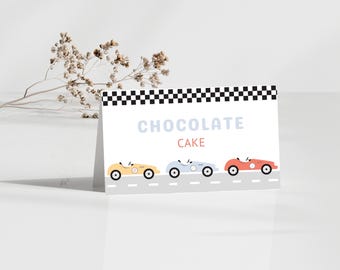 Peut inclure: Une carte blanche avec une bordure en damier noir et blanc. La carte indique "CHOCOLATE CAKE" en bleu et rouge. Il y a trois voitures de course dessinées en ligne sur la carte, une jaune, une bleue et une rouge.