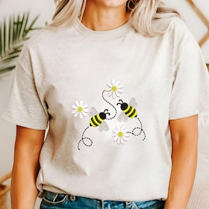以下が含まれることがあります： ベージュのTシャツ。黄色と黒のストライプの2匹の蜂が白いデイジーの近くを飛んでいる、気まぐれなデザイン。蜂は点線で花に繋がっており、遊び心のある陽気な美学を作り出しています。