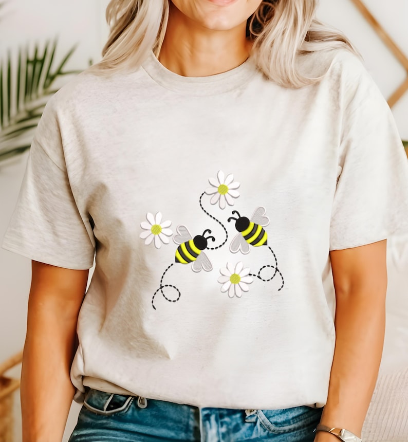 以下が含まれることがあります： ベージュのTシャツ。黄色と黒のストライプの2匹の蜂が白いデイジーの近くを飛んでいる、気まぐれなデザイン。蜂は点線で花に繋がっており、遊び心のある陽気な美学を作り出しています。