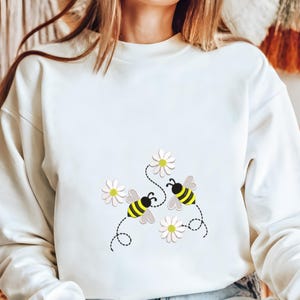 以下が含まれることがあります： クリーム色のスウェットシャツ。黄色と黒の縞模様の漫画のミツバチが白いデイジーの近くを飛んでいる、気まぐれなデザイン。ミツバチは点線で花に繋がっており、遊び心のある陽気な美学を作り出しています。