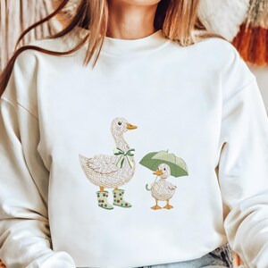 Może przedstawiać: Kremowa bluza z haftowanym wzorem matki kaczki w zielonej kokardzie i kaloszach oraz małej kaczki pod zielonym parasolem. Kaczki są w szczeg&oacute;łowym, teksturowanym stylu.