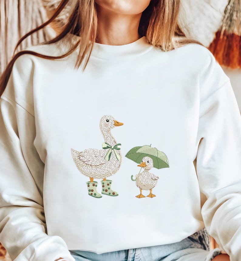 Może przedstawiać: Kremowa bluza z haftowanym wzorem matki kaczki w zielonej kokardzie i kaloszach oraz małej kaczki pod zielonym parasolem. Kaczki są w szczeg&oacute;łowym, teksturowanym stylu.