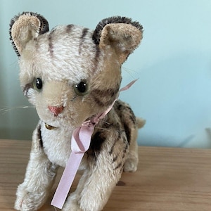 Puede incluir: Un juguete de gato de peluche vintage con un cuerpo de color beige claro y rayas marrones oscuras. El gato tiene una cinta rosa alrededor del cuello y una pequeña campana. El gato está sentado sobre una superficie de madera.
