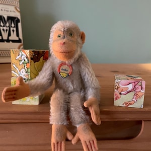 May include: A vintage stuffed monkey toy with grey fur, orange hands and feet, and a round orange face. The monkey has a red tag that says "Jocko". Two small, colourful blocks are on either side of the monkey.