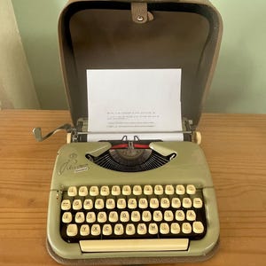 Könnte beinhalten: Eine tragbare Schreibmaschine in Salbeigrün mit QWERTZ-Tastaturlayout und einem braunen Kofferdeckel. Das Gerät verfügt über cremefarbene Tasten, einen Wagenrücklaufhebel aus Metall und ein eingelegtes Blatt weißes Papier mit gedrucktem Text.