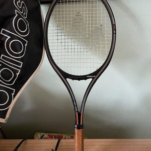 Könnte beinhalten: Ein Vintage Adidas Ivan Lendl Tennisschläger mit einem schwarzen und silbernen Rahmen, weißen Saiten und einem braunen Ledergriff. Der Schläger hat das Adidas-Logo und den Text "Ivan Lendl" auf dem Rahmen. Eine schwarze Adidas-Schlägertasche ist im Hintergrund zu sehen.