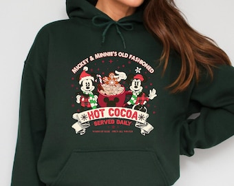 May include: Dark green hooded sweatshirt featuring a graphic of Mickey and Minnie Mouse serving hot cocoa. The graphic includes the text "Mickey & Minnie's Old Fashioned Hot Cocoa Served Daily" in a festive design. The sweatshirt has a front pocket and a drawstring hood.