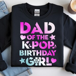 May include: Black crewneck sweatshirt with the text "DAD of the KPOP BIRTHDAY GIRL" in pink, white, and blue gradient lettering. The sweatshirt is next to a pair of blue jeans and a cup of coffee.