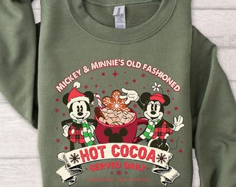 May include: An olive green sweatshirt featuring a graphic of Mickey and Minnie Mouse in holiday attire, with the text "Mickey & Minnie's Old Fashioned Hot Cocoa Served Daily". The design includes a cocoa mug and gingerbread cookies.