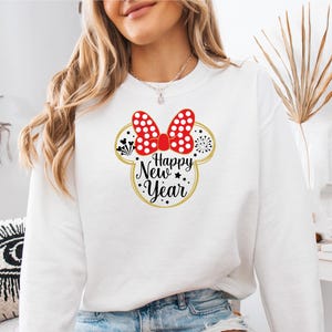 May include: White sweatshirt featuring a Minnie Mouse design with a red polka dot bow and the text "Happy New Year" in black and gold. The design includes fireworks and stars, perfect for celebrating the new year.