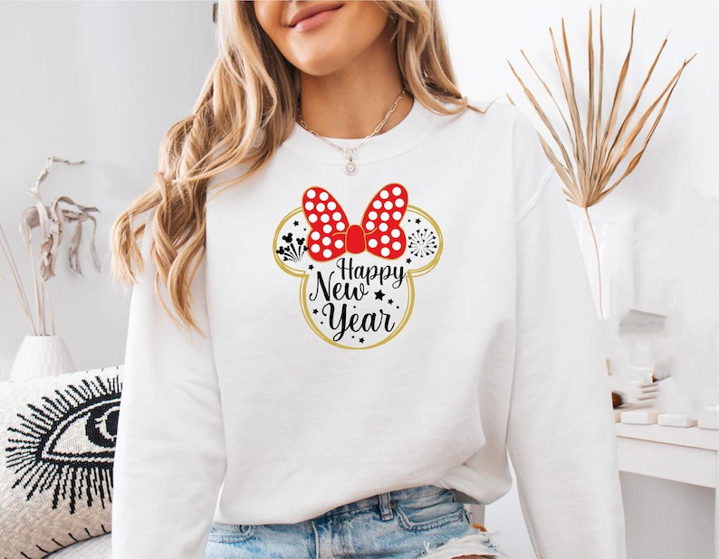 May include: White sweatshirt featuring a Minnie Mouse design with a red polka dot bow and the text "Happy New Year" in black and gold. The design includes fireworks and stars, perfect for celebrating the new year.