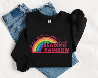 May include: Black sweatshirt with a rainbow and the words "Take a Look, it's in a Book! READING RAINBOW" in a distressed font. The sweatshirt is folded on top of a pair of blue jeans.