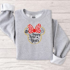 May include: A light gray sweatshirt with rolled-up sleeves. The front features a Minnie Mouse-inspired design with a red bow with white polka dots, a gold outline, and the words "Happy New Year" in black script.