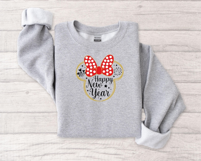 May include: A light gray sweatshirt with rolled-up sleeves. The front features a Minnie Mouse-inspired design with a red bow with white polka dots, a gold outline, and the words "Happy New Year" in black script.