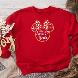 May include: A red sweatshirt featuring a Minnie Mouse-inspired design with a white polka dot bow and the words "Happy New Year" in a gold outline. The design includes stars and snowflakes. The sweatshirt is part of a holiday collection.