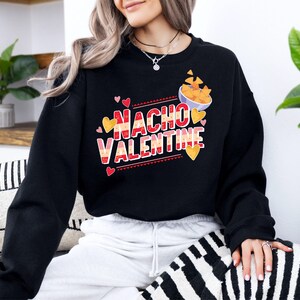 May include: Black sweatshirt with the text "Nacho Valentine" in red and white, with a bowl of nachos and heart graphics. The sweatshirt is a casual style, perfect for Valentine's Day or a fun, food-themed gift.