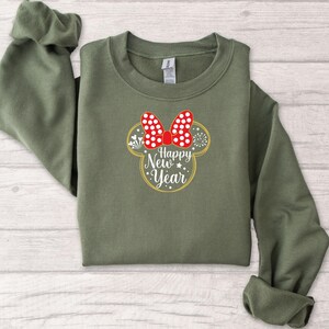 May include: An olive green sweatshirt featuring a Minnie Mouse-inspired design with a red bow with white polka dots. The design includes the text "Happy New Year" in a gold outline, with stars and fireworks accents.