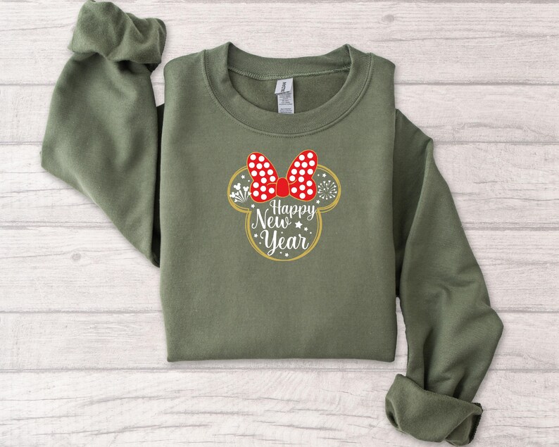 May include: An olive green sweatshirt featuring a Minnie Mouse-inspired design with a red bow with white polka dots. The design includes the text "Happy New Year" in a gold outline, with stars and fireworks accents.