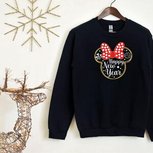 May include: Black sweatshirt with a Minnie Mouse-inspired design. The design features a red bow with white polka dots, a gold outline, and the words "Happy New Year" in white. The sweatshirt is hanging on a wooden hanger.