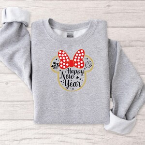 May include: A light gray sweatshirt with rolled-up sleeves. The front features a Minnie Mouse-inspired design with a red polka-dot bow, fireworks, and the words "Happy New Year" in a gold outline.