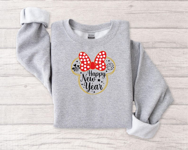 May include: A light gray sweatshirt with rolled-up sleeves. The front features a Minnie Mouse-inspired design with a red polka-dot bow, fireworks, and the words "Happy New Year" in a gold outline.