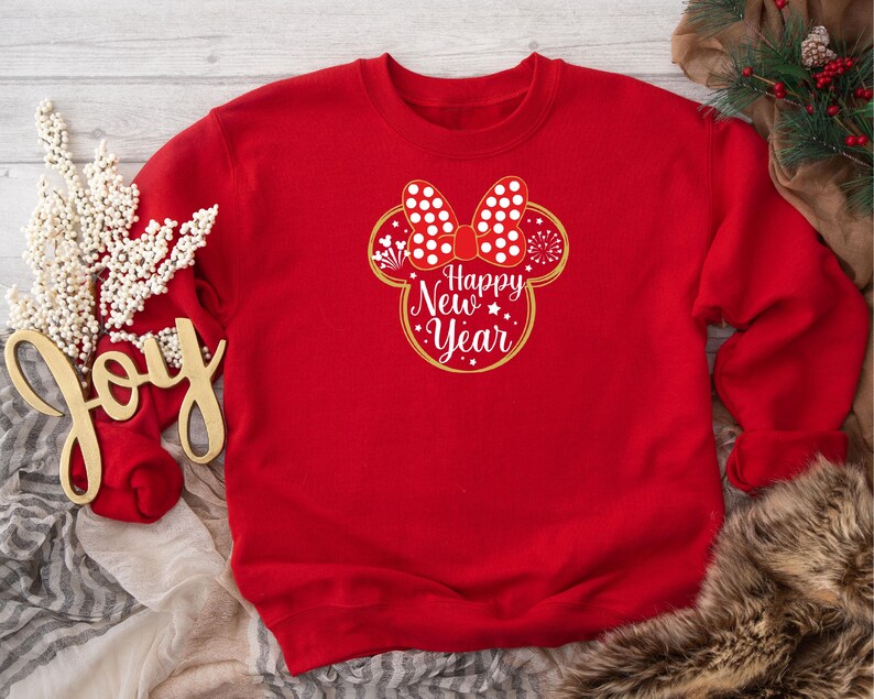 May include: A red sweatshirt featuring a Minnie Mouse-inspired design with a white polka dot bow and the words "Happy New Year" in a gold outline. The sweatshirt is part of a holiday collection.
