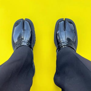 Könnte beinhalten: Ein Paar schwarze Schuhe mit geteilter Zehenpartie und glänzender Oberfläche, vor einem leuchtend gelben Hintergrund. Die Schuhe haben ein einzigartiges Design mit einem separaten Fach für den großen Zeh. Die Schuhe werden mit einer schwarzen Hose getragen.
