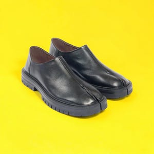 Könnte beinhalten: Ein Paar schwarze Leder-Slipper mit geteiltem Zehendesign. Die Schuhe haben eine dicke Profilsohle und einen flachen Absatz. Die Schuhe werden auf einem leuchtend gelben Hintergrund präsentiert.