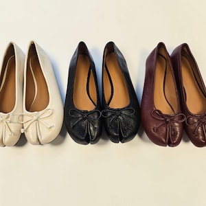 May include: A collection of three pairs of ballet flats in cream, black, and burgundy. Each pair features a rounded toe, a small bow detail, and a split-toe design. The shoes are arranged in a row against a white background.
