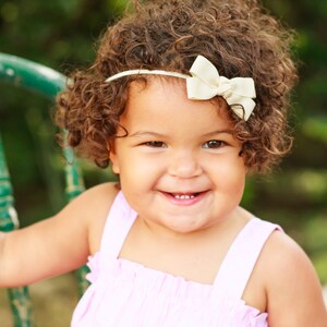 May include: A young girl with curly brown hair is wearing a pink dress and a white headband with a bow. She is smiling and looking at the camera.