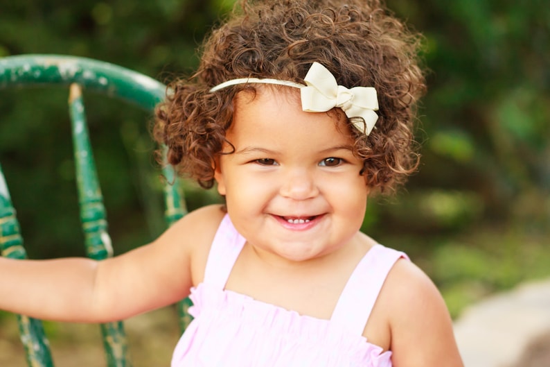 May include: A young girl with curly brown hair is wearing a pink dress and a white headband with a bow. She is smiling and looking at the camera.