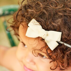 May include: A young girl with curly brown hair wearing a white bow headband.