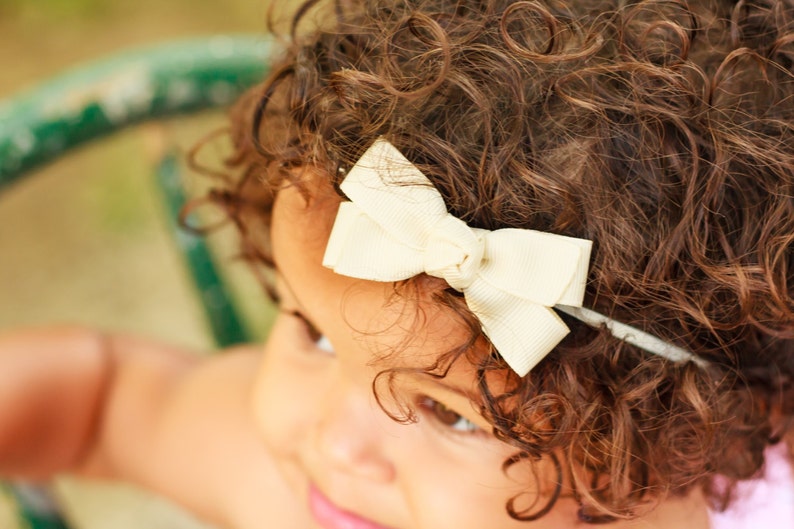 May include: A young girl with curly brown hair wearing a white bow headband.