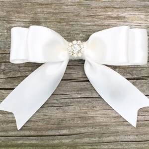 May include: A white satin bow with a clear rhinestone center. The bow is tied with a simple knot and the ends are left long.