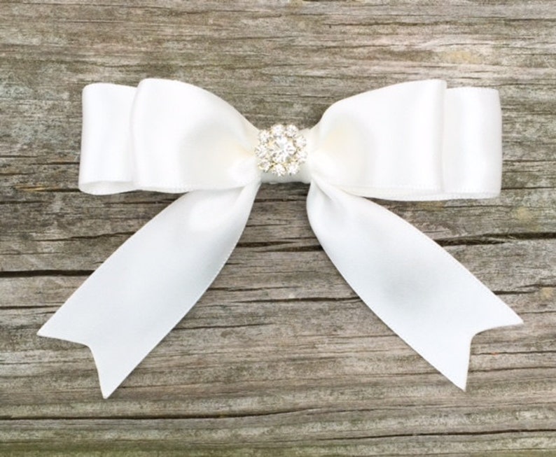 May include: A white satin bow with a clear rhinestone center. The bow is tied with a simple knot and the ends are left long.
