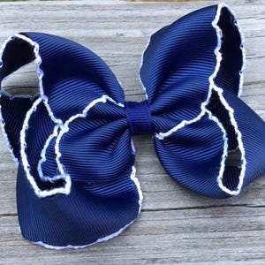 May include: A navy blue hair bow with white scalloped edges. The bow is made of grosgrain ribbon and is tied in a classic bow shape.
