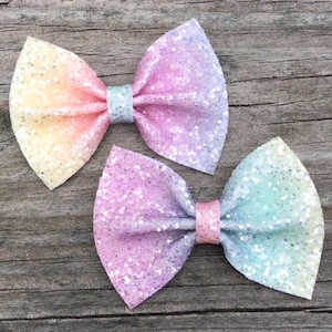May include: Two pastel ombre glitter hair bows. The bows are tied with a silver ribbon and have a gradient of pink, yellow, and blue.