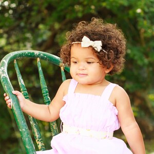 May include: A young girl with curly brown hair wearing a pink dress with a white belt and a white bow headband sits on a green wooden chair.