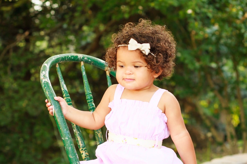 May include: A young girl with curly brown hair wearing a pink dress with a white belt and a white bow headband sits on a green wooden chair.