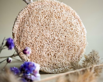 Op de afbeelding: Een natuurlijke loofah spons met een lusvormig handvat. De spons is lichtbruin en heeft een ruwe textuur. Hij ligt op een tafel met gedroogde bloemen en grassen.