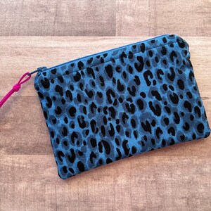 May include: A rectangular blue zippered pouch with a black leopard print pattern. The pouch has a pink cord zipper pull. The pouch is made of fabric and is laying on a wooden surface.