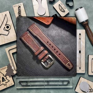 May include: A brown leather watch strap with white stitching and a silver buckle rests on a textured black surface. Wooden craft tools and leather pieces are arranged around the strap, indicating a leathercraft theme.