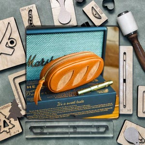 May include: An orange leather pencil case with a leaf design, gold zipper, and pull tab. The case rests on a blue box with gold text, next to a gold pen. Various wooden craft tools and stamps surround the case.
