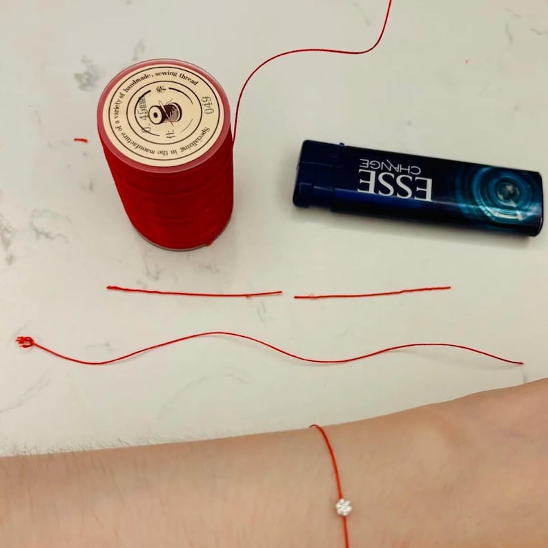 May include: A red string bracelet with a small clear stone charm. The bracelet is on a person's wrist. There is a spool of red thread, a lighter, and two pieces of red string on a white surface.