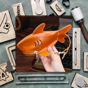 Puede incluir: Un monedero de cuero naranja con forma de tiburón con cierre de cremallera y un asa pequeña. El bolso tiene una aleta en la parte superior y una serie de tres líneas diagonales en el lateral. El fondo incluye herramientas de artesanía en cuero y sellos de madera.