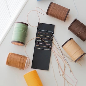 May include: A variety of spools of thread in different colors, including green, brown, beige, and orange. The spools are arranged on a white surface with a black leather swatch showing different stitching patterns.